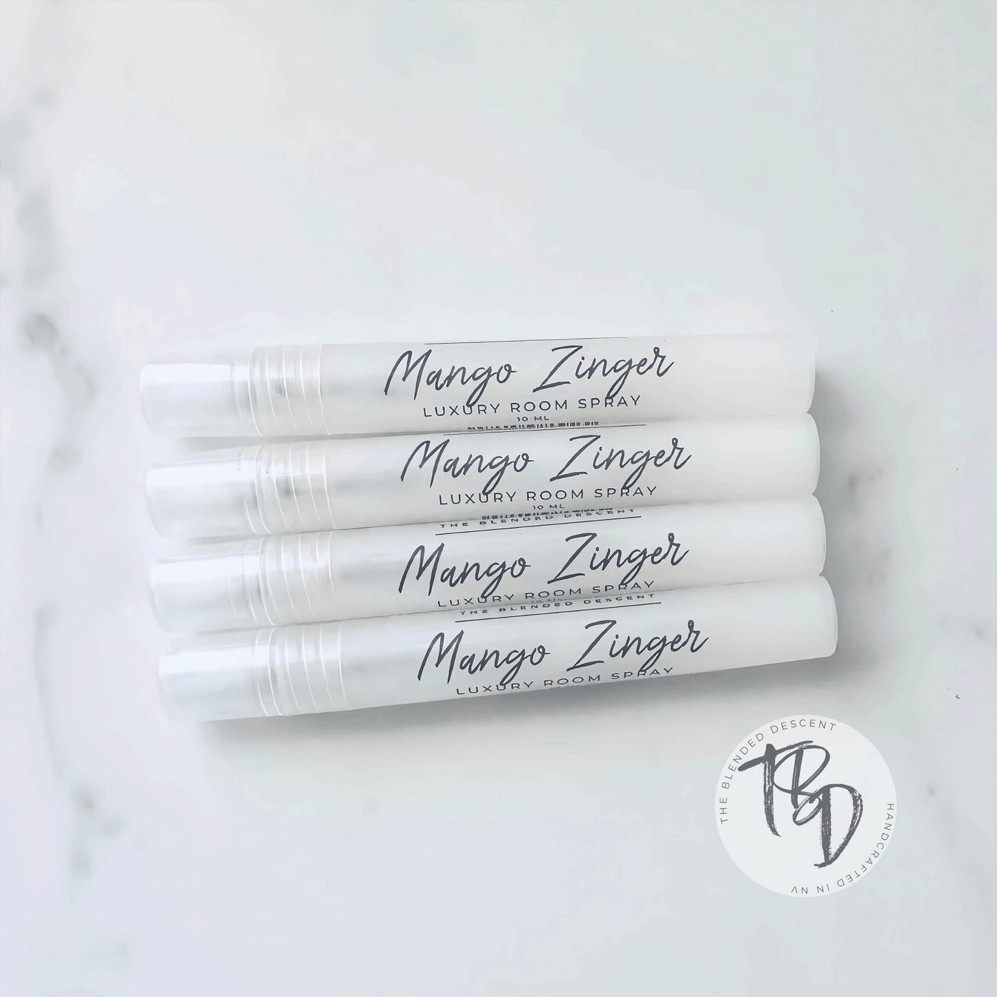 Four white spray bottles labeled 'Mango Zinger Luxury Room Spray' arranged in a stack on a light surface. Each bottle has a sleek, minimalist design with black cursive text, and a small circular logo in the bottom right corner of the image