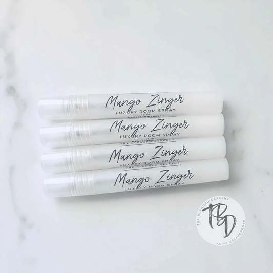 Four white spray bottles labeled 'Mango Zinger Luxury Room Spray' arranged in a stack on a light surface. Each bottle has a sleek, minimalist design with black cursive text, and a small circular logo in the bottom right corner of the image