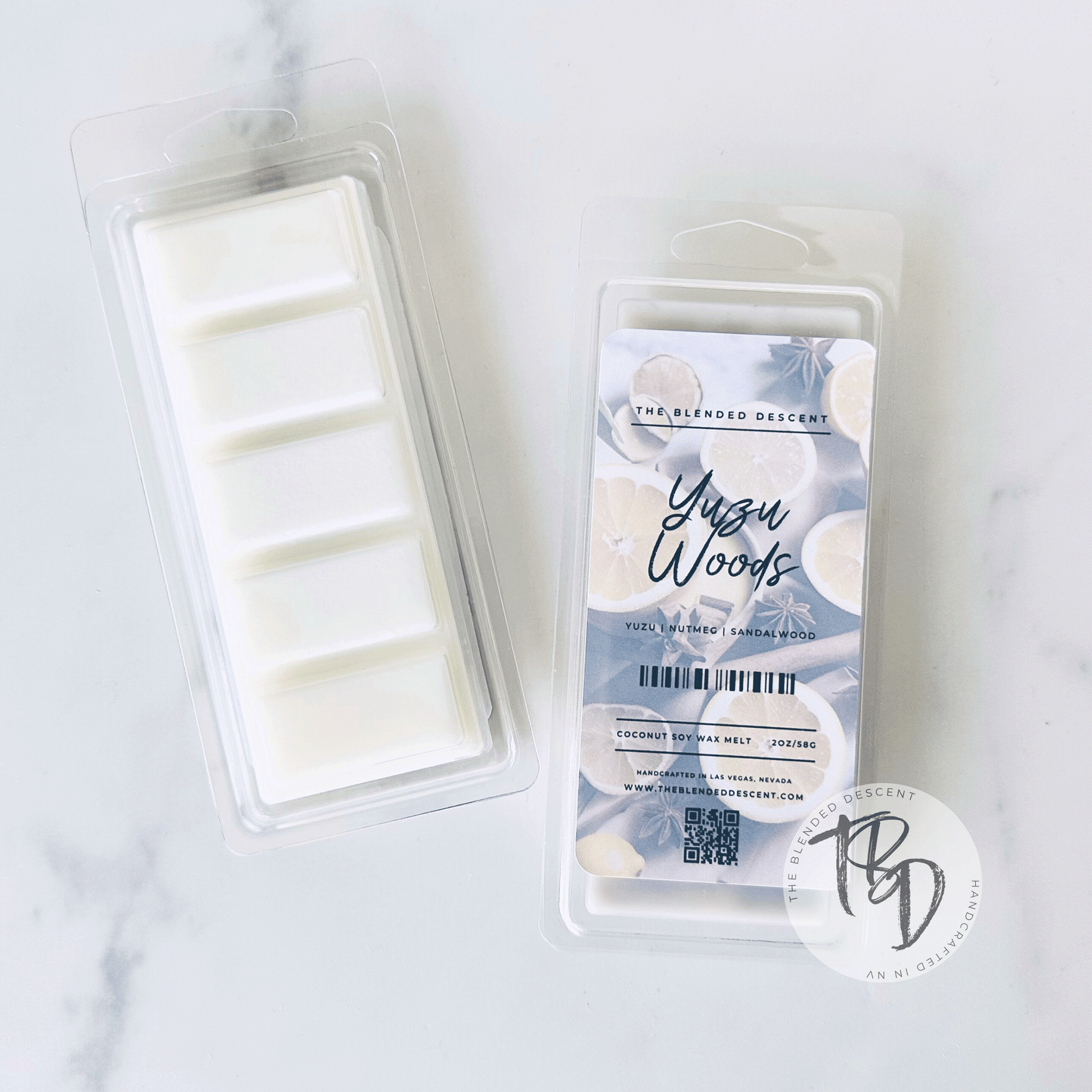 The image shows a package of wax melts with the product label "Yuzu Woods" from "The Blended Descent." The packaging highlights key fragrance notes such as yuzu, nutmeg, and sandalwood. The front of the label features images of lemon slices, star anise, and cinnamon sticks, emphasizing the citrusy and spicy notes. It is marked as a "Coconut Soy Wax Melt," weighing 2 oz/56g. The product was handcrafted in Las Vegas, Nevada, with additional information available via a QR code on the packaging.